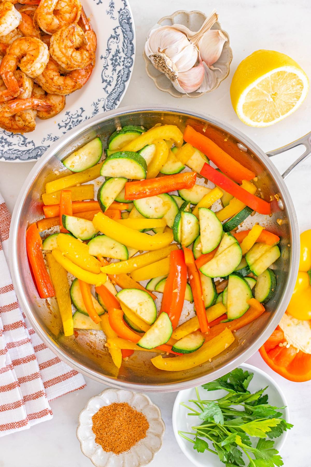 Sautéed bell peppers and zucchini in a pan, surrounded by shrimp, lemon, garlic, parsley, and spices.
