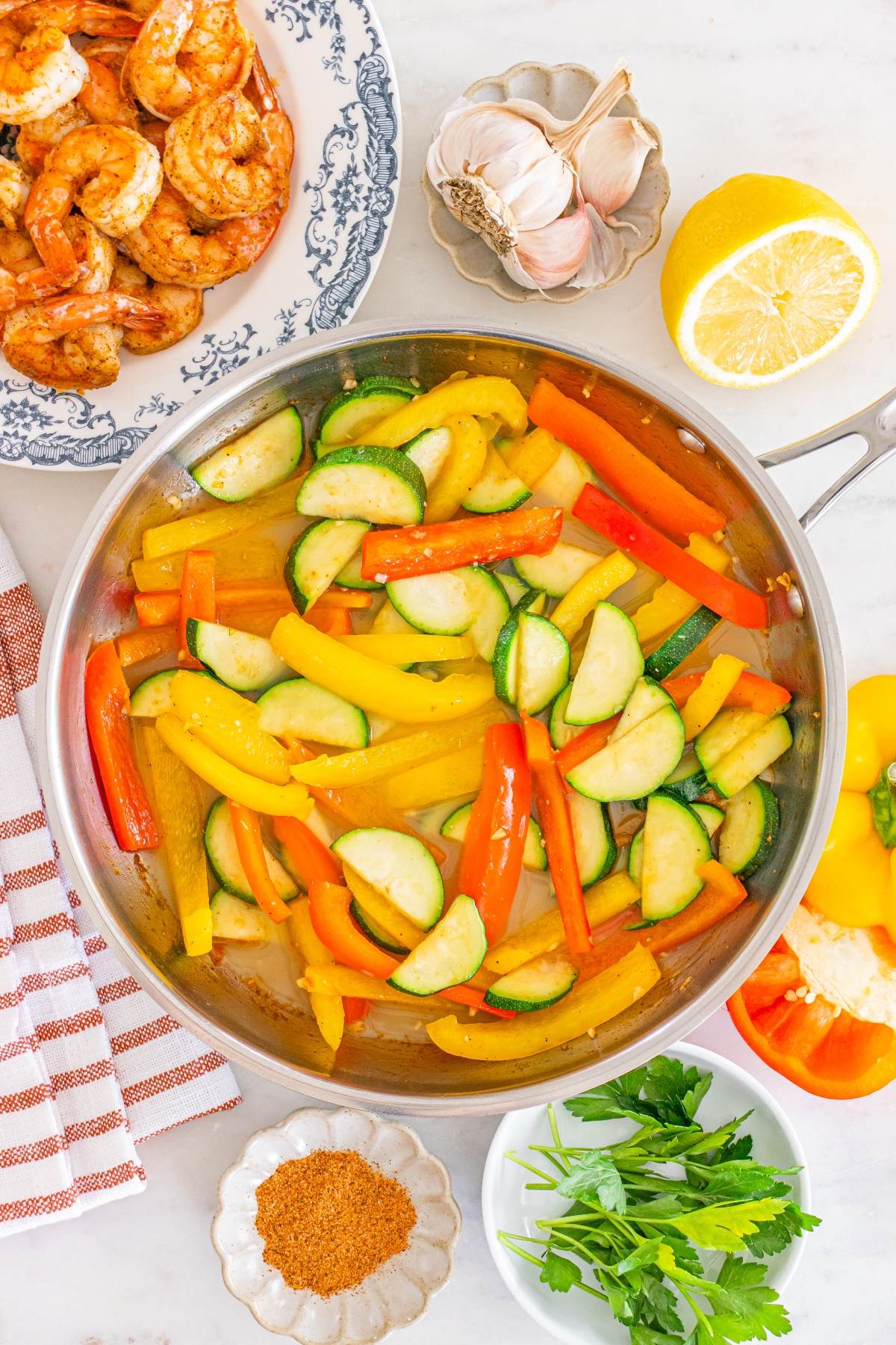 Colorful sautéed bell peppers and zucchini in a pan, surrounded by shrimp, spices, parsley, garlic, and lemon.