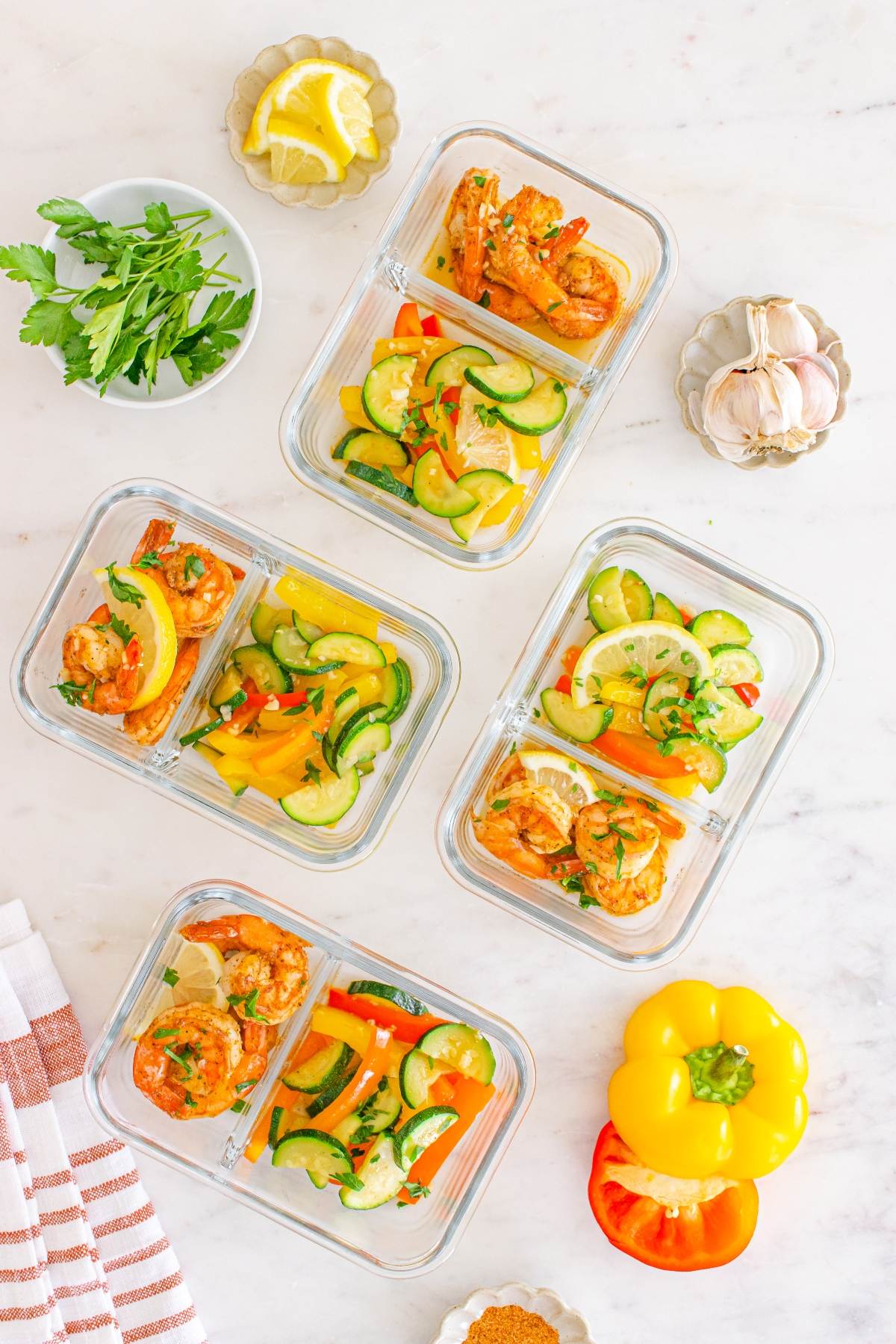 Four glass meal prep containers with shrimp, zucchini, and bell pepper on a marble surface with fresh ingredients.