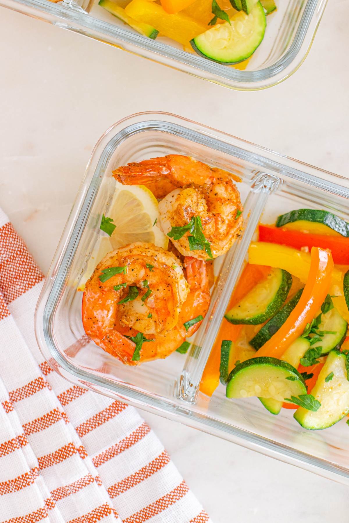 Glass container with cooked shrimp, lemon slices, and mixed sautéed vegetables, on a striped towel.