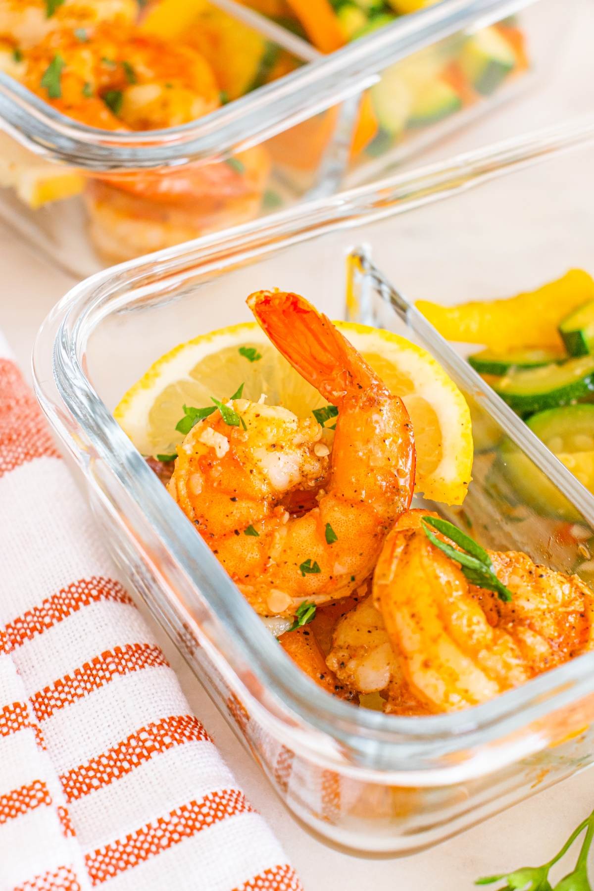 Glass container with cooked shrimp, lemon slices, and vegetables, next to a red-and-white striped towel.