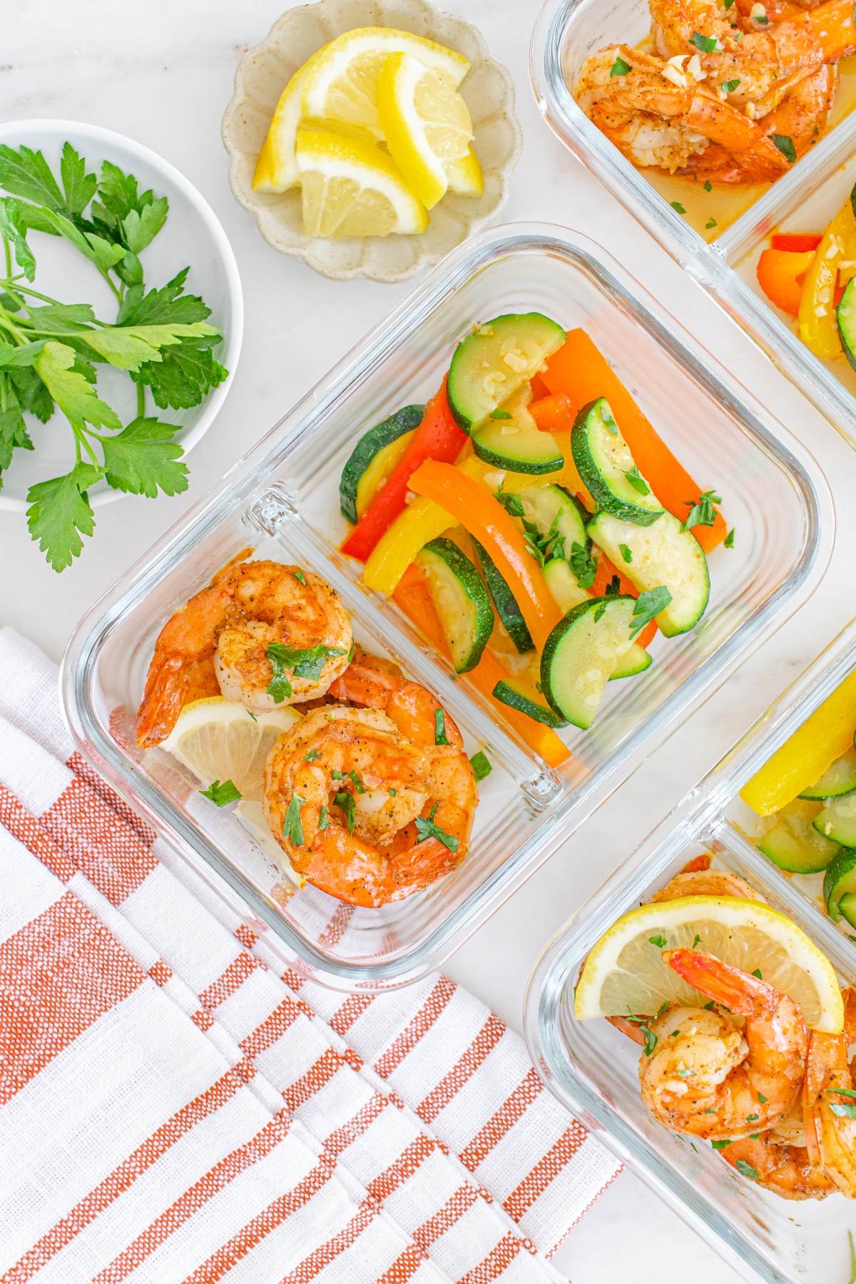 Glass meal prep containers with shrimp, lemon slices, and sautéed zucchini and bell peppers on a white surface.