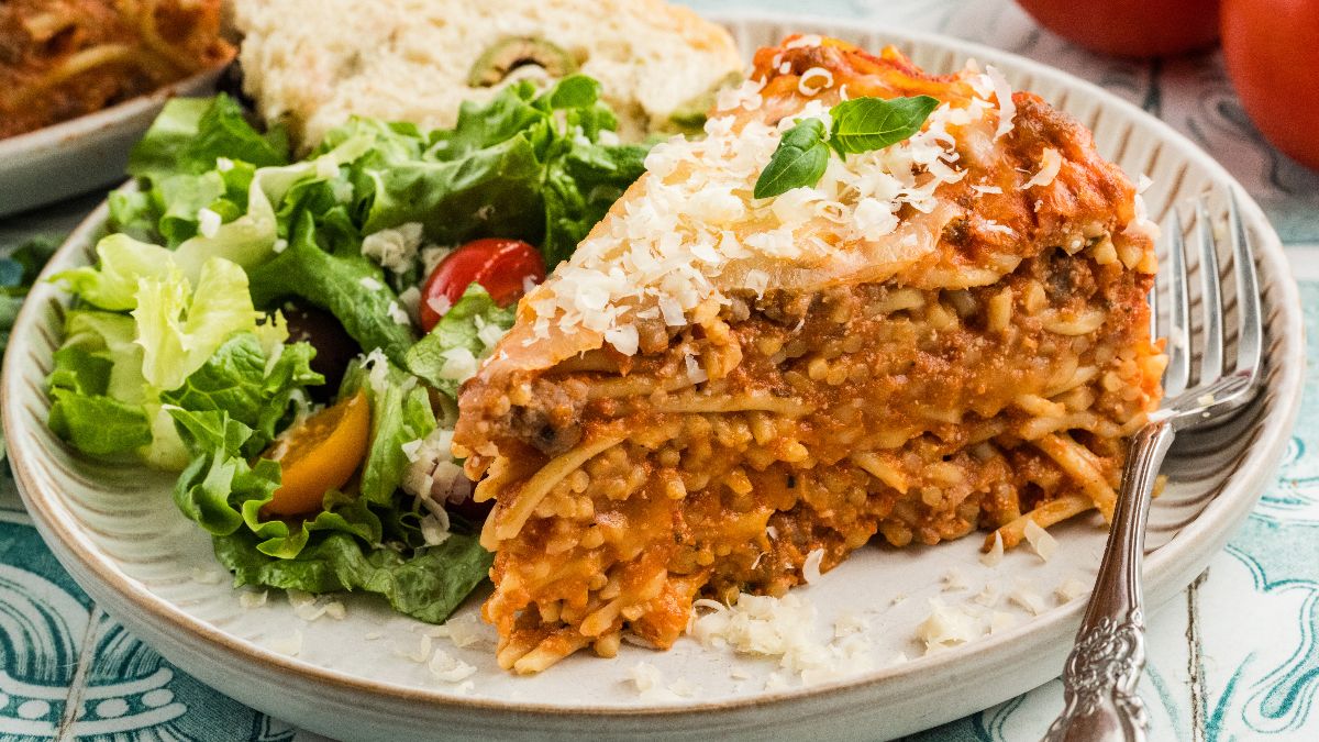 Slice of spaghetti pie with cheese and basil, served with salad and bread on a plate.