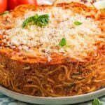 A delicious spaghetti pie crowned with melted cheese and fragrant basil rests on a plate, encircled by fresh vegetables and a splash of vibrant pesto.