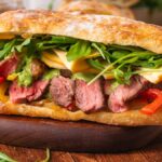 Steak Sandwich with sliced steak, melty cheese, arugula, grilled peppers, and savory sauce on fresh ciabatta bread.