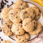 Banana chocolate chip cookies rest invitingly on a plate, surrounded by fresh bananas, scattered chocolate chips, and a striped cloth.