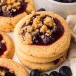 Blueberry pie cookies adorned with blueberry jam and a crumble topping, nestled among fresh blueberries on a light cloth.