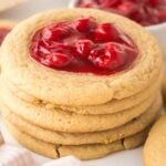 Stack of cherry thumbprint cookies with red cherry filling, on a light surface with a pink cloth nearby.