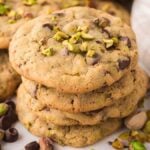 A tempting stack of pistachio chocolate chip cookies sits surrounded by scattered nuts and chocolate chips, enticing every sweet tooth.