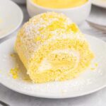 A slice of lemon swiss roll with cream filling, dusted with powdered sugar, sits on a white plate—a tempting treat from any classic lemon roll cake recipe.