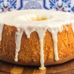 A lemon chiffon cake glazed with white icing sits on a wooden board, with fresh lemons and strawberries in the background.