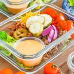 Glass container with lettuce, ground beef, cherry tomatoes, pickles, onions, cheese, bacon, and a sauce cup—perfect for tasty meal prep bacon cheeseburger bowls.