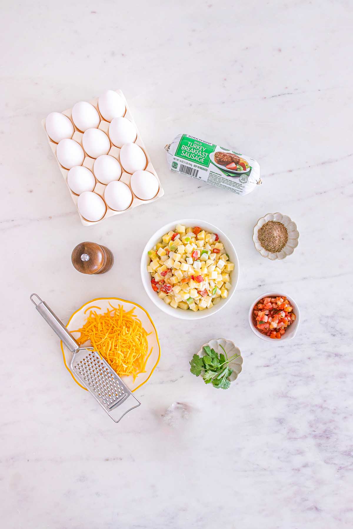 Eggs, shredded cheese, potatoes, seasonings, pico de gallo, sausage, and cilantro arranged on a white surface for easy meal prep breakfast taco bowls.