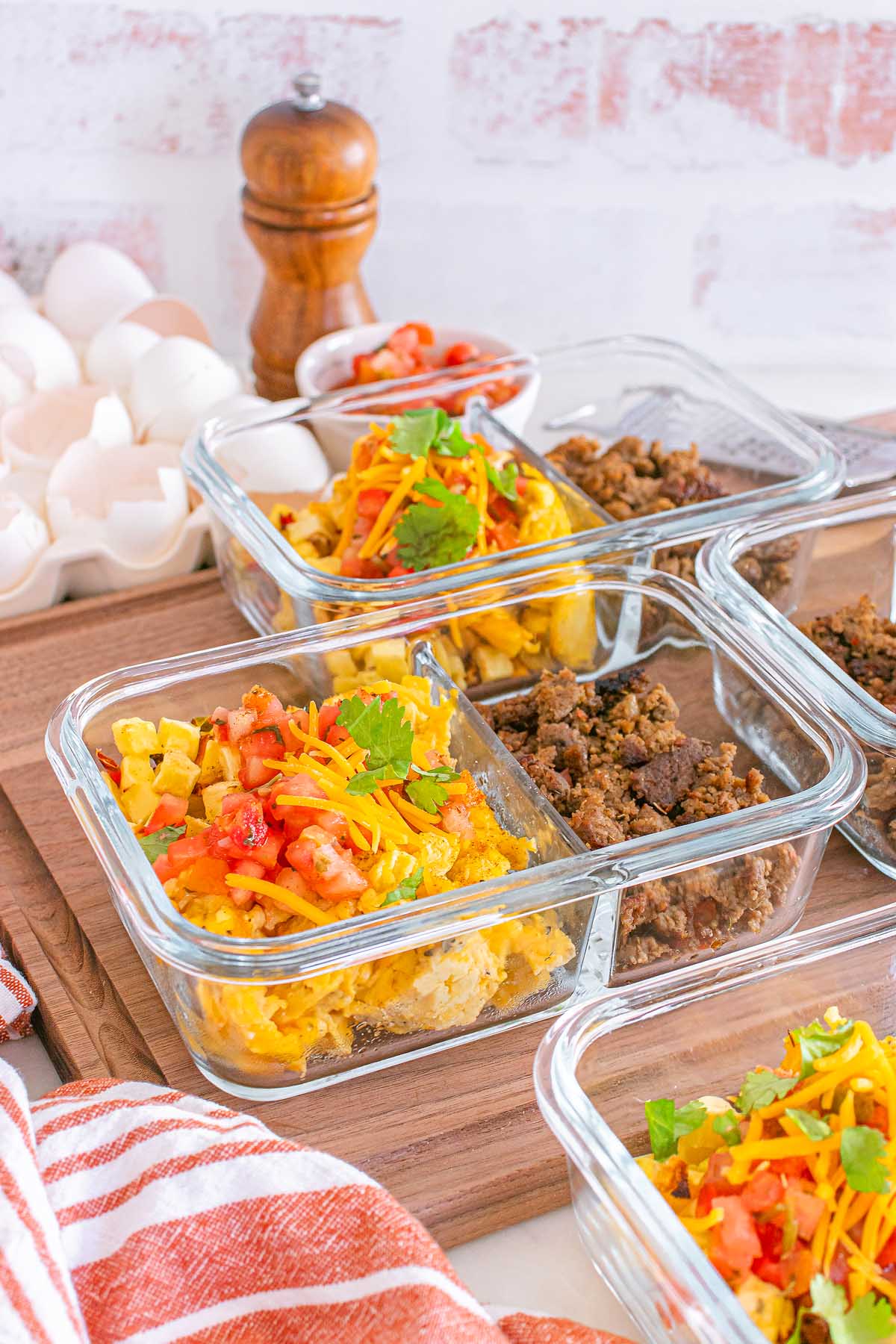 Glass containers with colorful meal prep breakfast taco bowls, featuring eggs, ground meat, veggies, and fresh herbs on a wooden board.