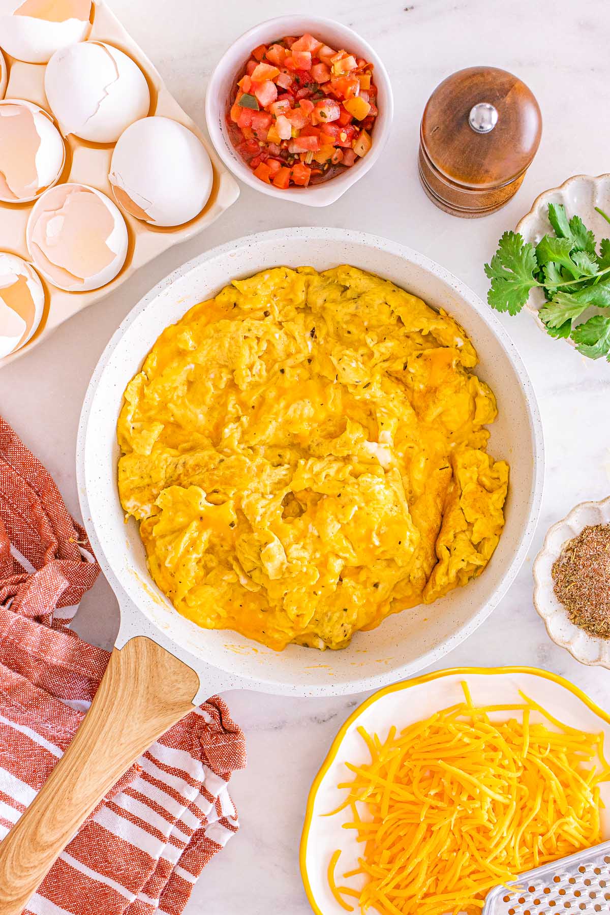 Scrambled eggs in a skillet with shredded cheese, salsa, eggshells, and herbs on a white countertop—perfect for meal prep breakfast taco bowls.