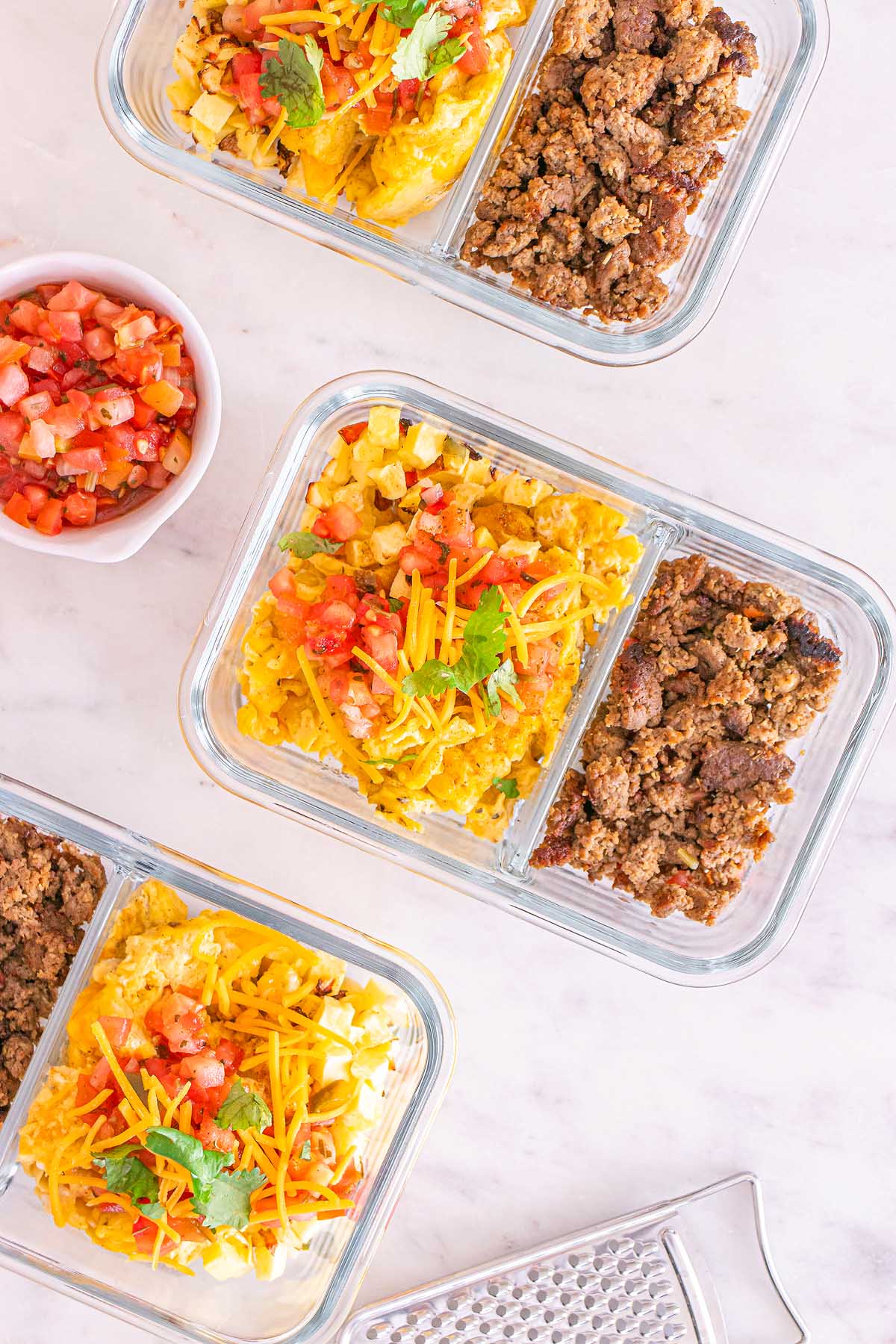 Glass meal prep containers filled with seasoned ground beef, shredded cheese, eggs, and salsa create convenient and delicious meal prep breakfast taco bowls on a white surface.
