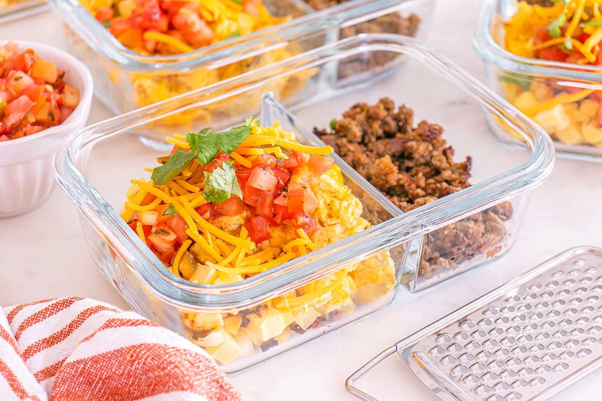 Glass meal prep container with breakfast taco bowls, featuring ground meat, shredded cheese, and diced tomatoes, next to a grater.