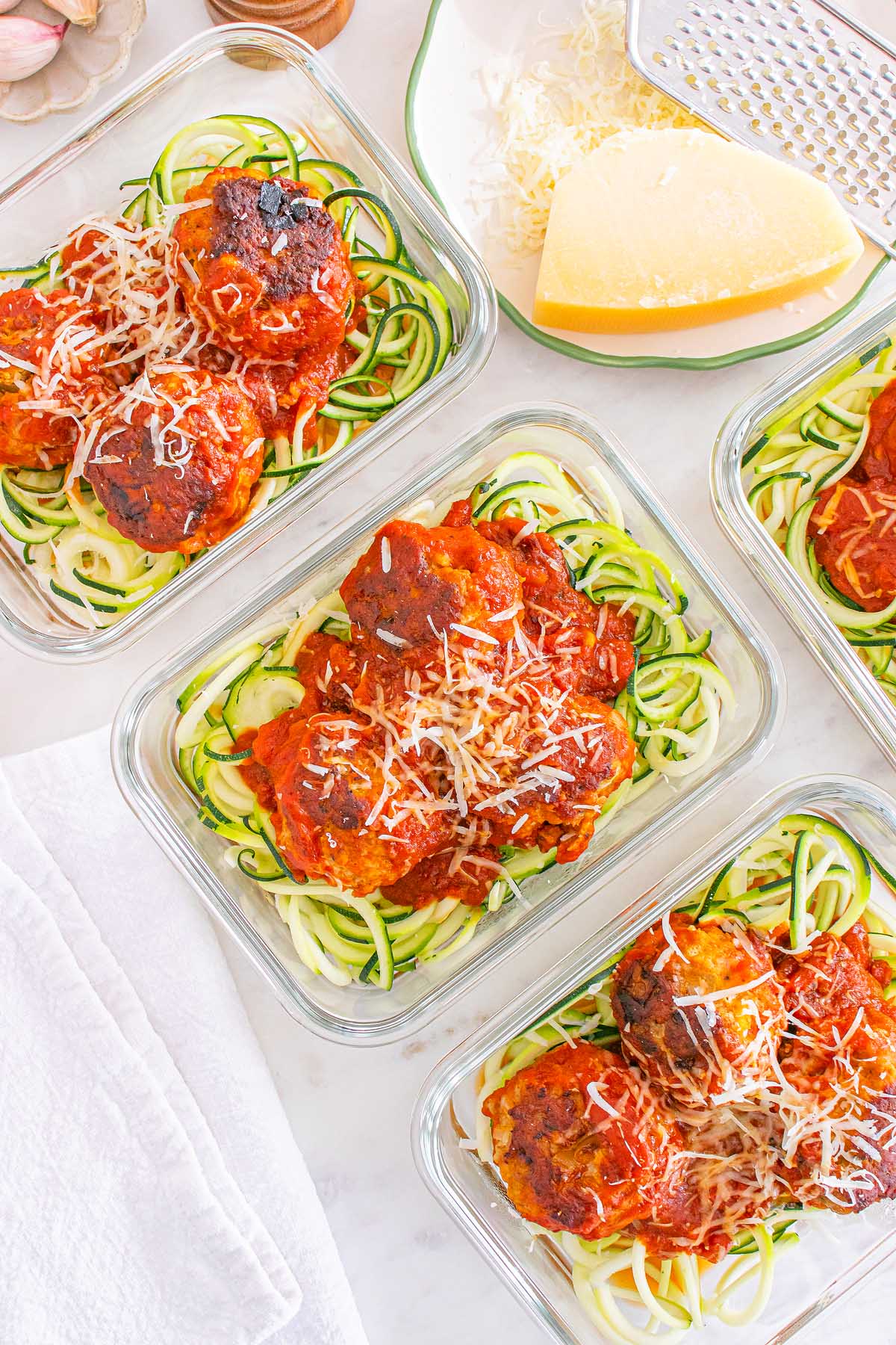 Glass containers with Meal Prep Turkey Meatballs & Zoodles in tomato sauce, topped with shredded cheese, next to a cheese wedge.