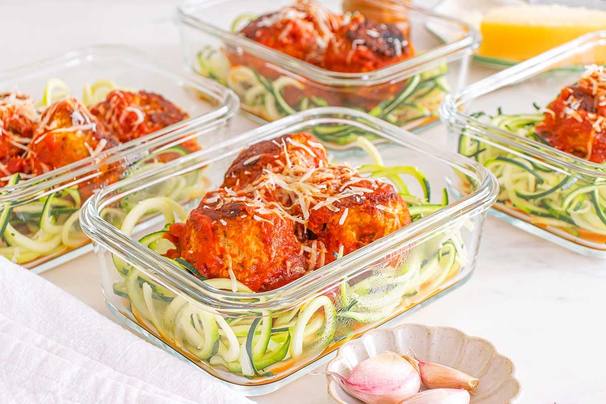 Glass containers with Meal Prep Turkey Meatballs & Zoodles in tomato sauce, topped with grated cheese, on a white surface.
