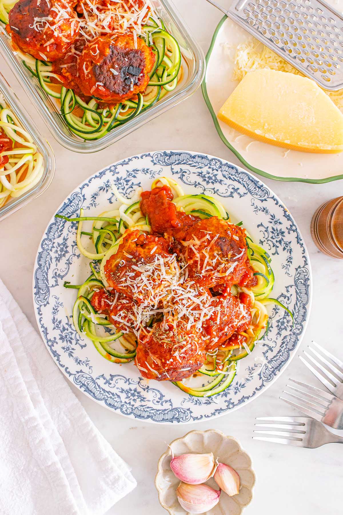 A plate of Meal Prep Turkey Meatballs & Zoodles in tomato sauce, topped with grated cheese, sits next to fresh cheese and garlic.