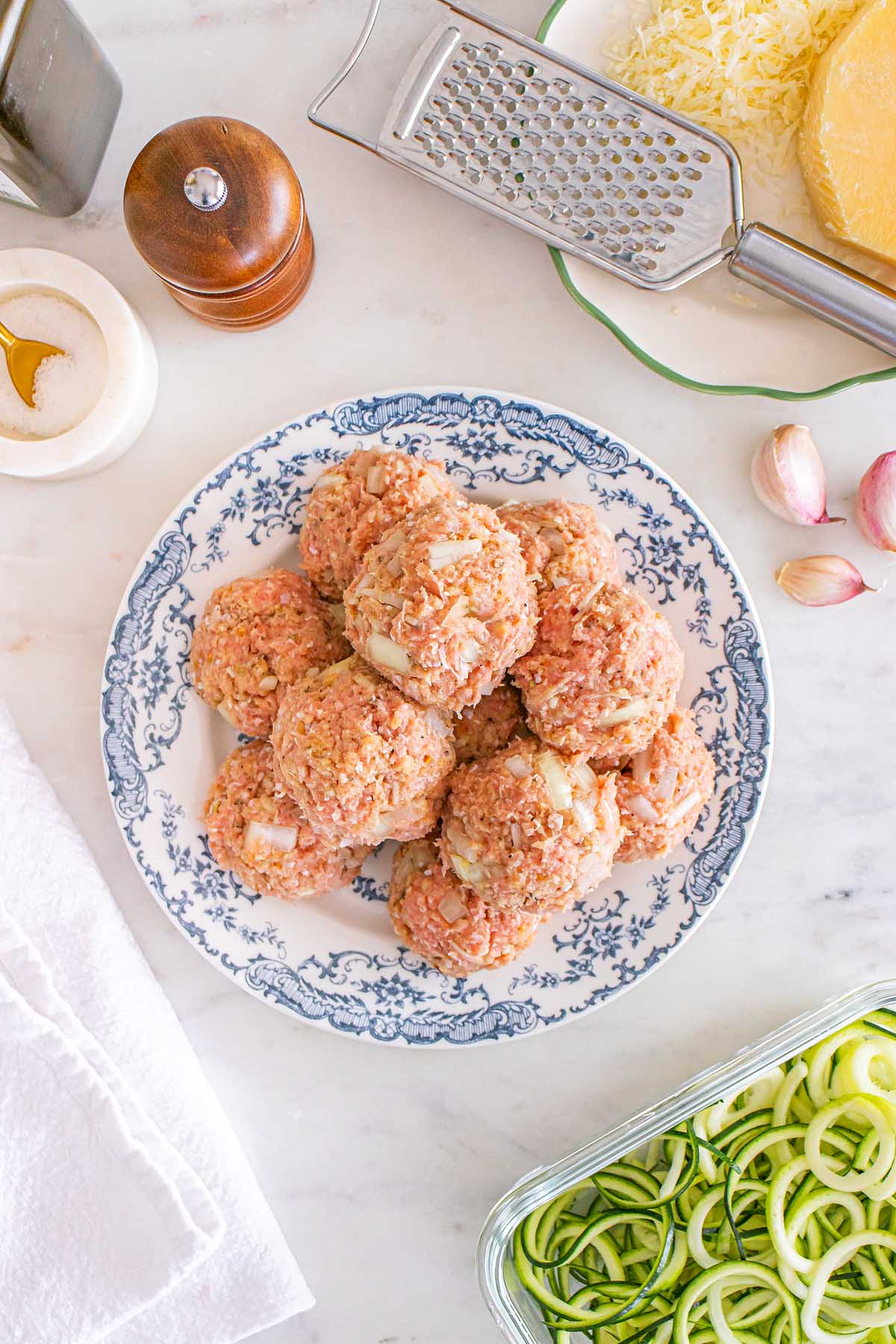 A plate of uncooked Meal Prep Turkey Meatballs on a blue and white dish, surrounded by cheese, garlic, and spiralized zucchini zoodles.