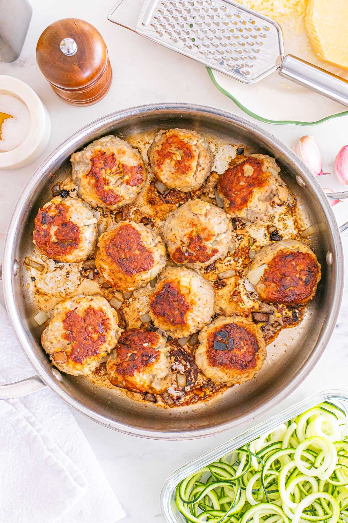 A pan of Meal Prep Turkey Meatballs browning on the stove, with fresh ingredients like cheese and zucchini zoodles nearby.
