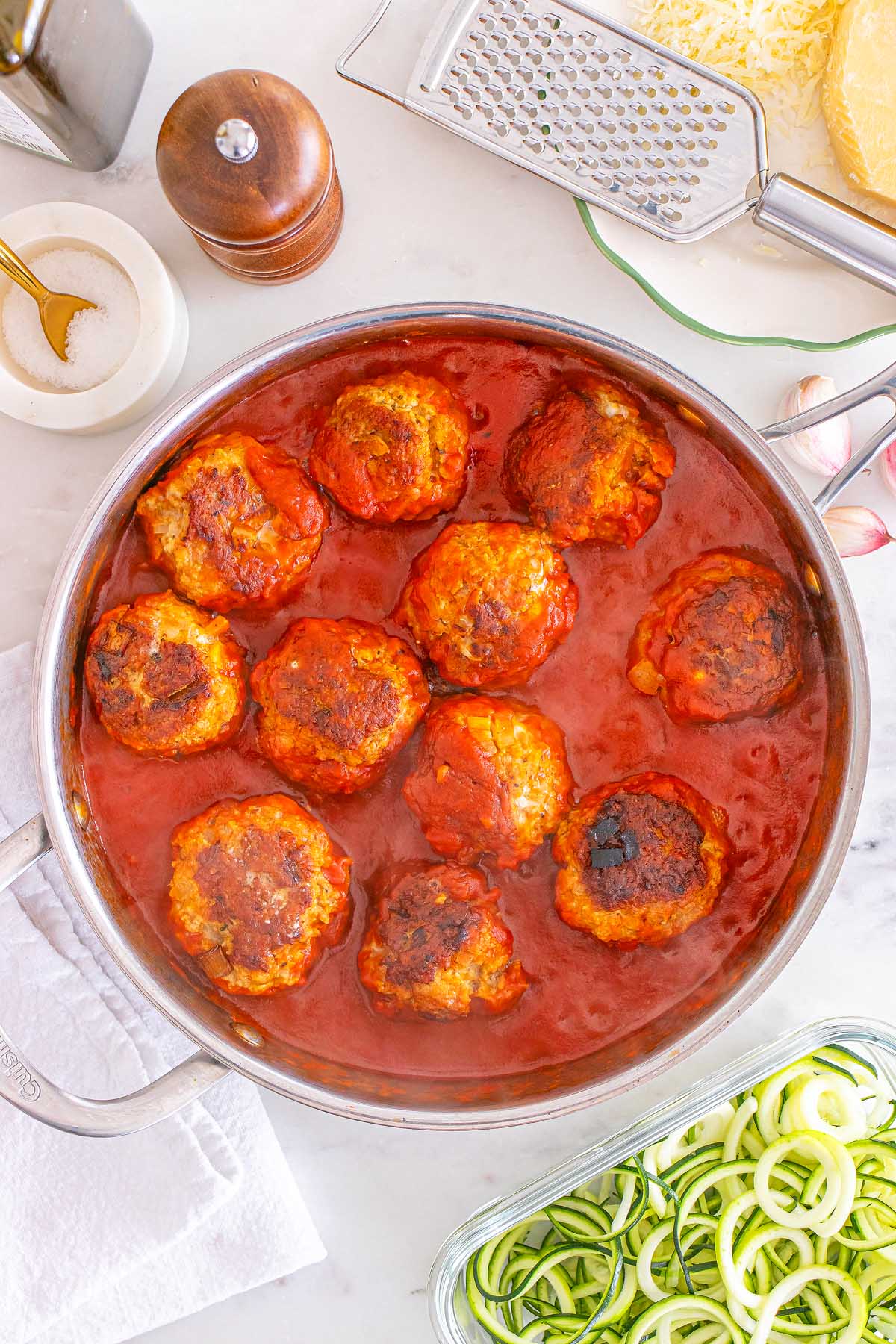 Meal Prep Turkey Meatballs simmer in tomato sauce in a pan, with grated cheese and spiralized zucchini zoodles nearby.