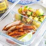 Glass container with meal prep Thai chicken slices, a lime wedge, and cucumber salad with red onion and herbs.