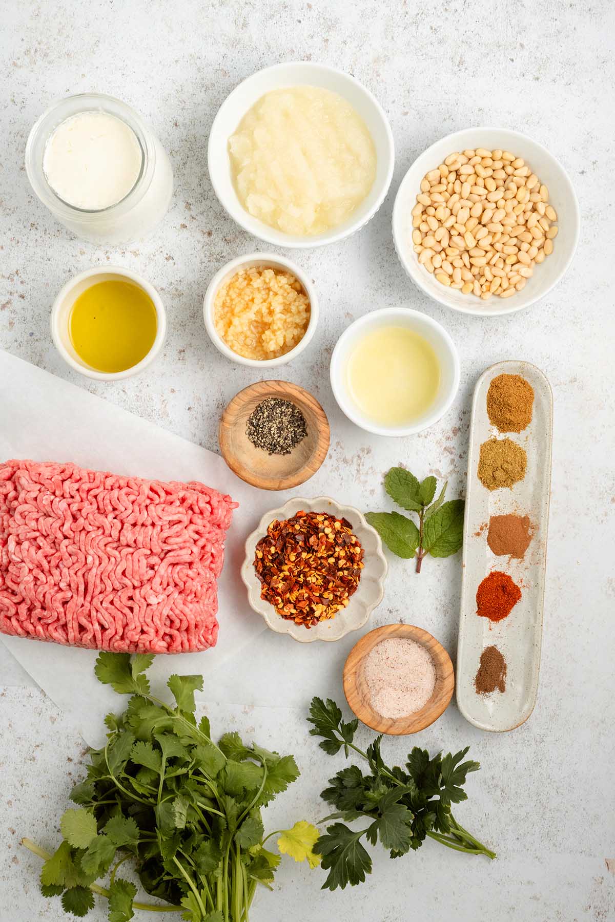 Ground beef, fresh herbs, spices, beans, oil, garlic, and sauces arranged neatly on a white surface&mdash;perfect for crafting flavorful beef kofta.