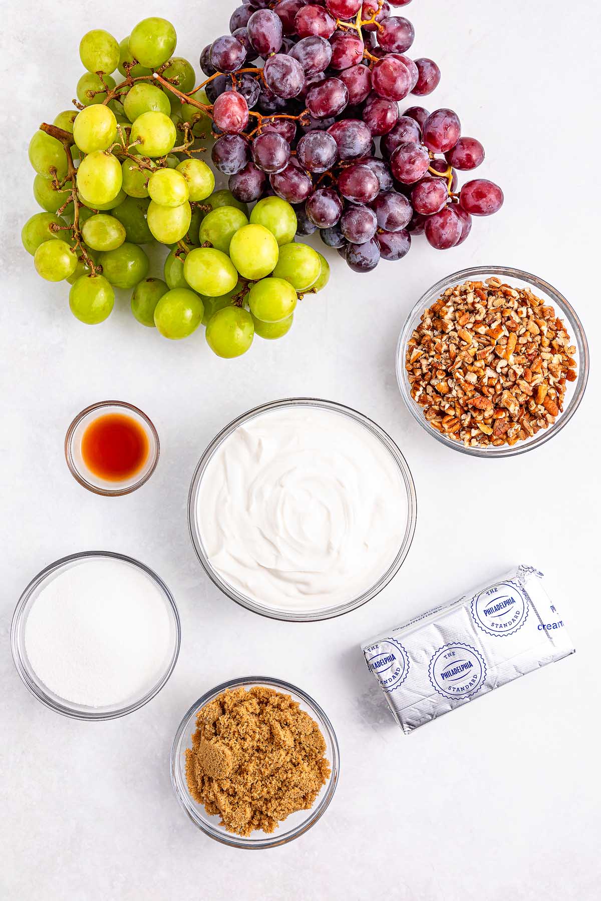 Grape salad featuring green and red grapes, pecans, cream cheese, yogurt, brown sugar, white sugar, and vanilla on a white surface.
