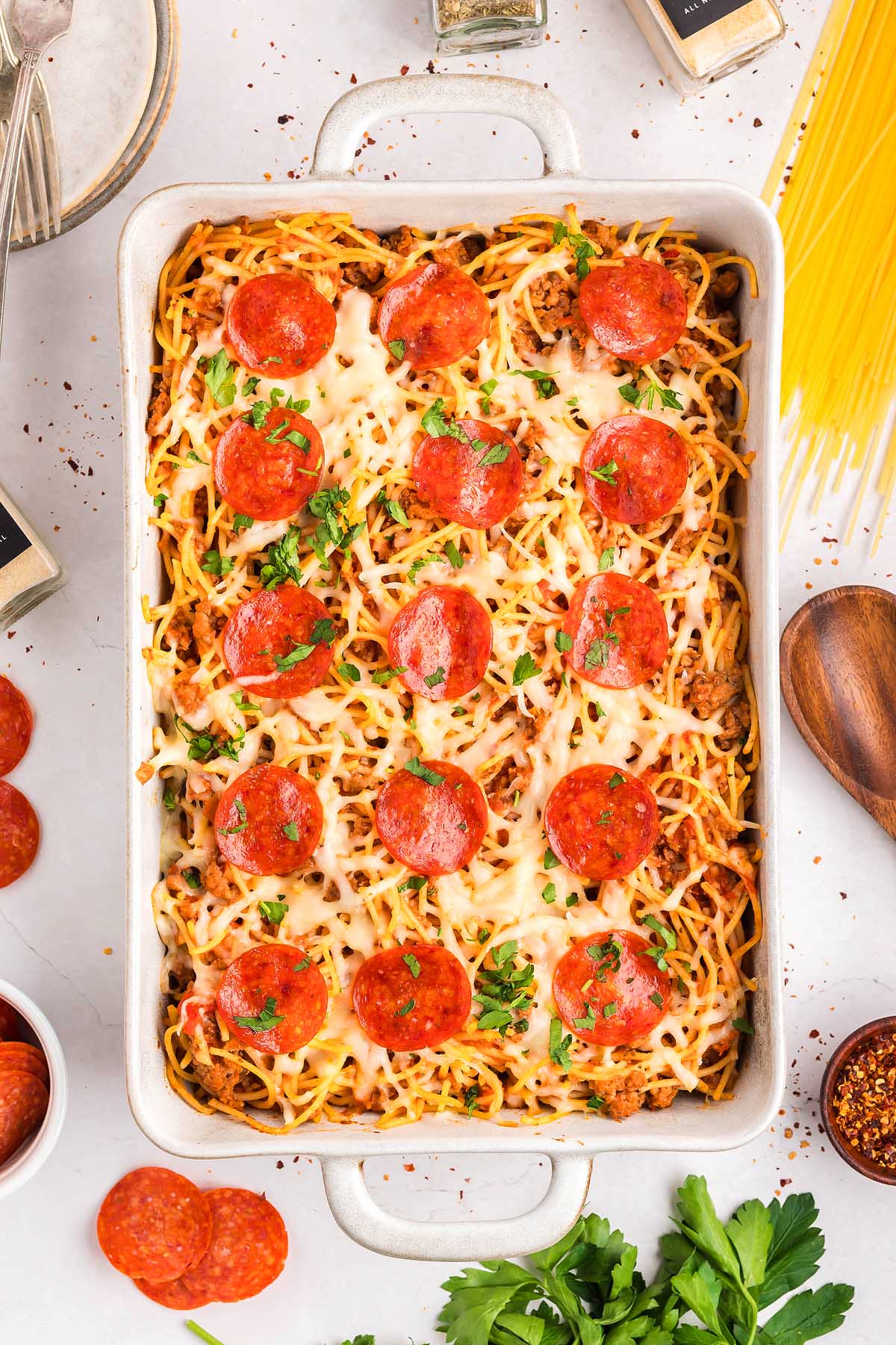 A casserole dish of baked spaghetti pizza topped with melted cheese, pepperoni slices, and chopped parsley.