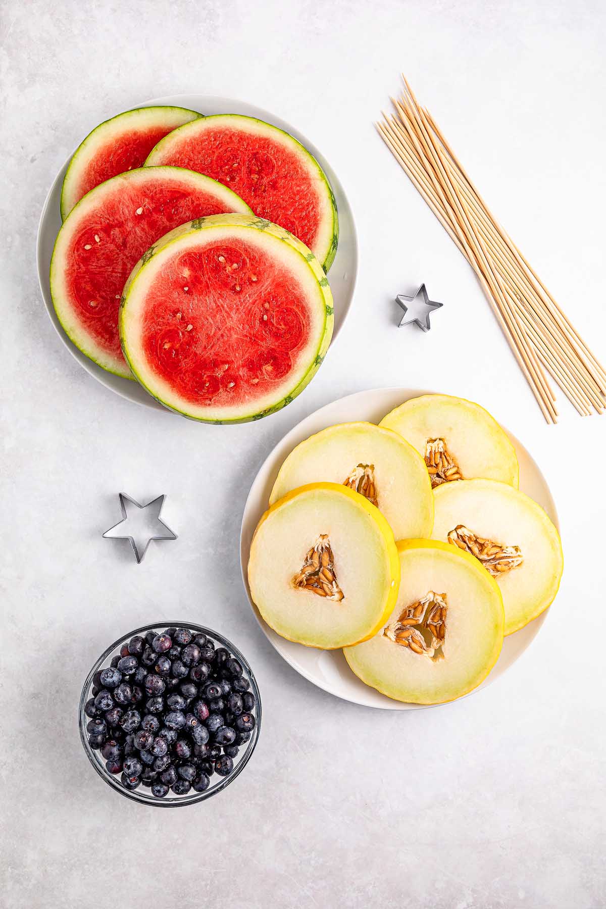 Plates of watermelon and melon slices, a bowl of blueberries, wooden skewers, and star-shaped cutters for making festive Watermelon Blueberry Sparklers.