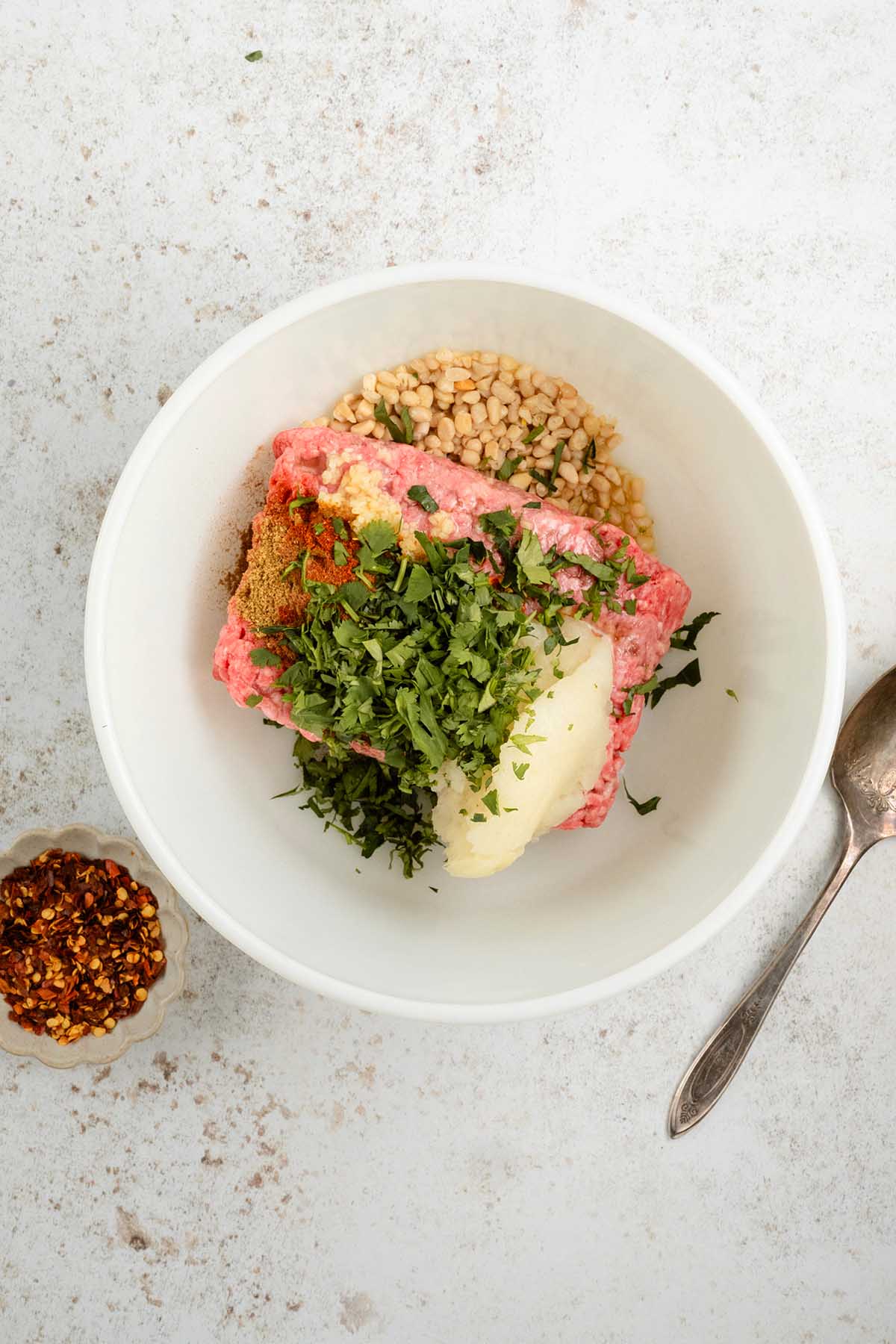 White bowl with ground meat for beef kofta, chopped herbs, pine nuts, and spices on a white surface; spoon and chili flakes nearby.