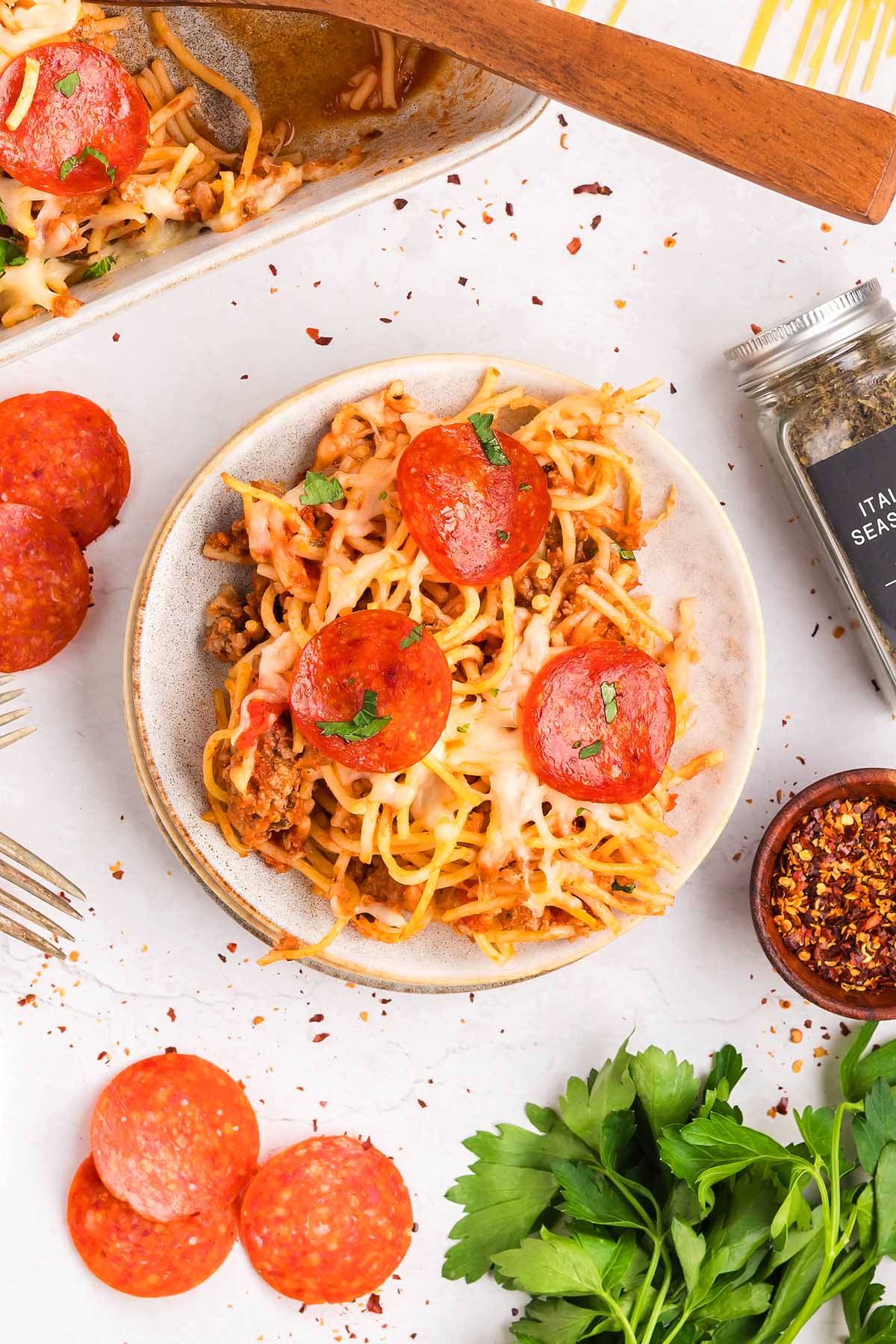A plate of baked spaghetti pizza topped with melted cheese and pepperoni, surrounded by herbs and spices.