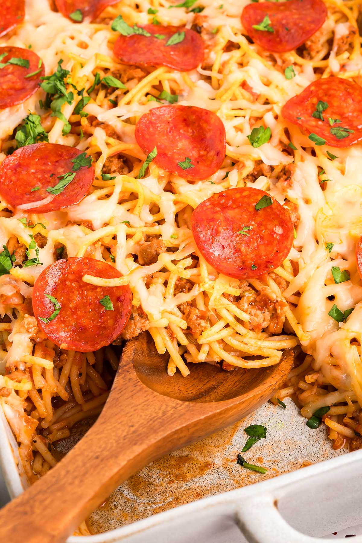 Baked spaghetti pizza topped with melted cheese, pepperoni slices, and parsley, served with a wooden spoon.
