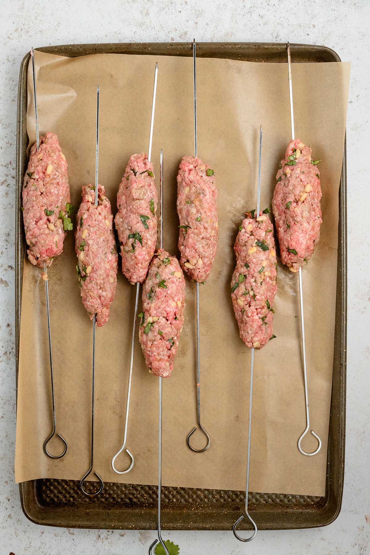 Six metal skewers with raw beef kofta mixture rest on a parchment-lined baking sheet, ready to be cooked.