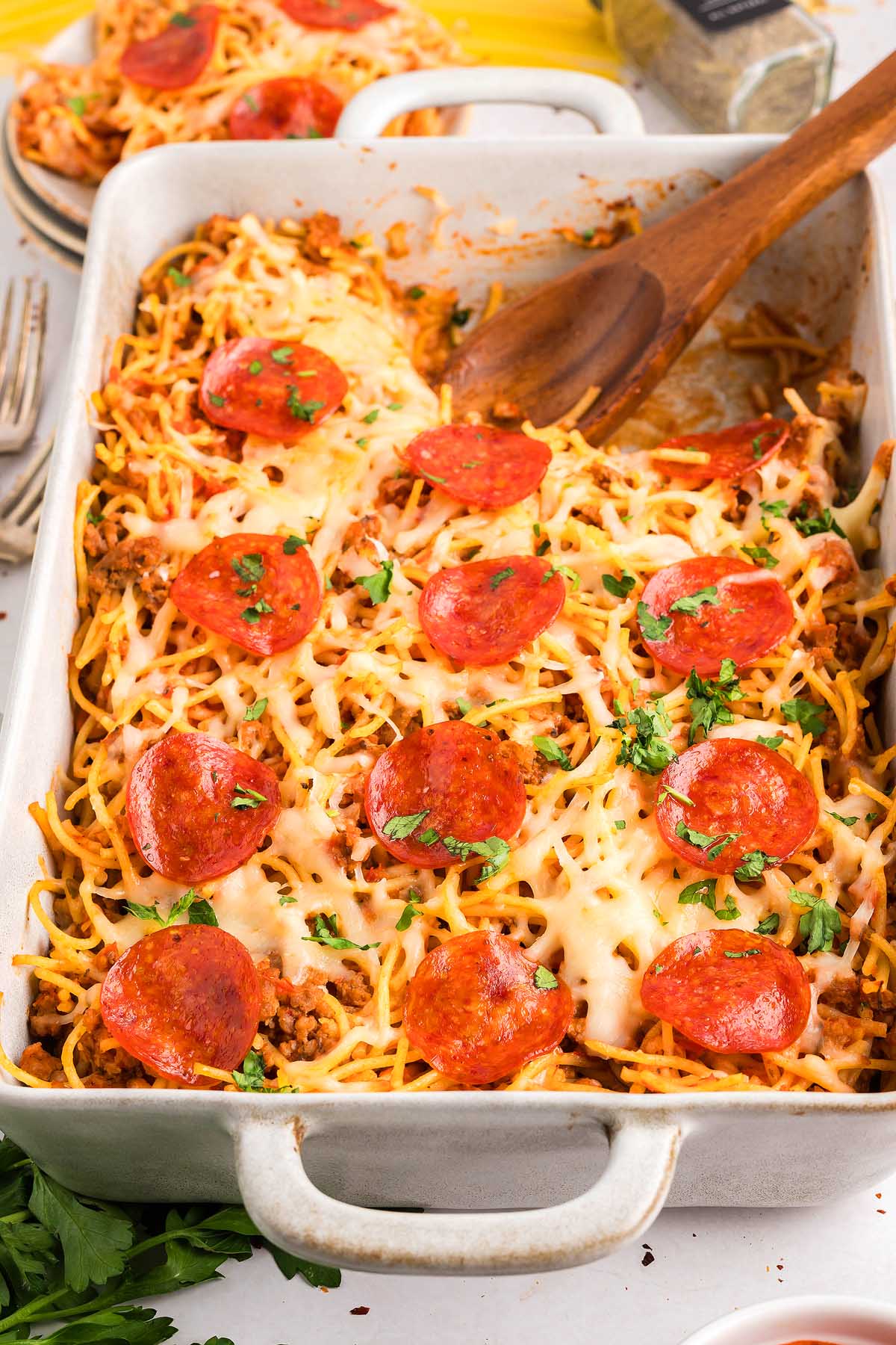 Baked spaghetti pizza casserole topped with melted cheese, pepperoni slices, and chopped parsley in a white dish.