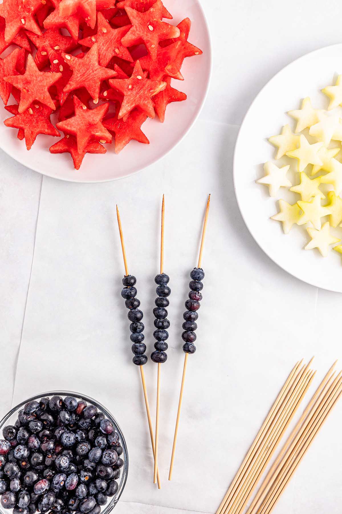 Three skewers with blueberries, next to plates of star-shaped Watermelon Blueberry Sparklers and melon slices, with a few loose skewers nearby.