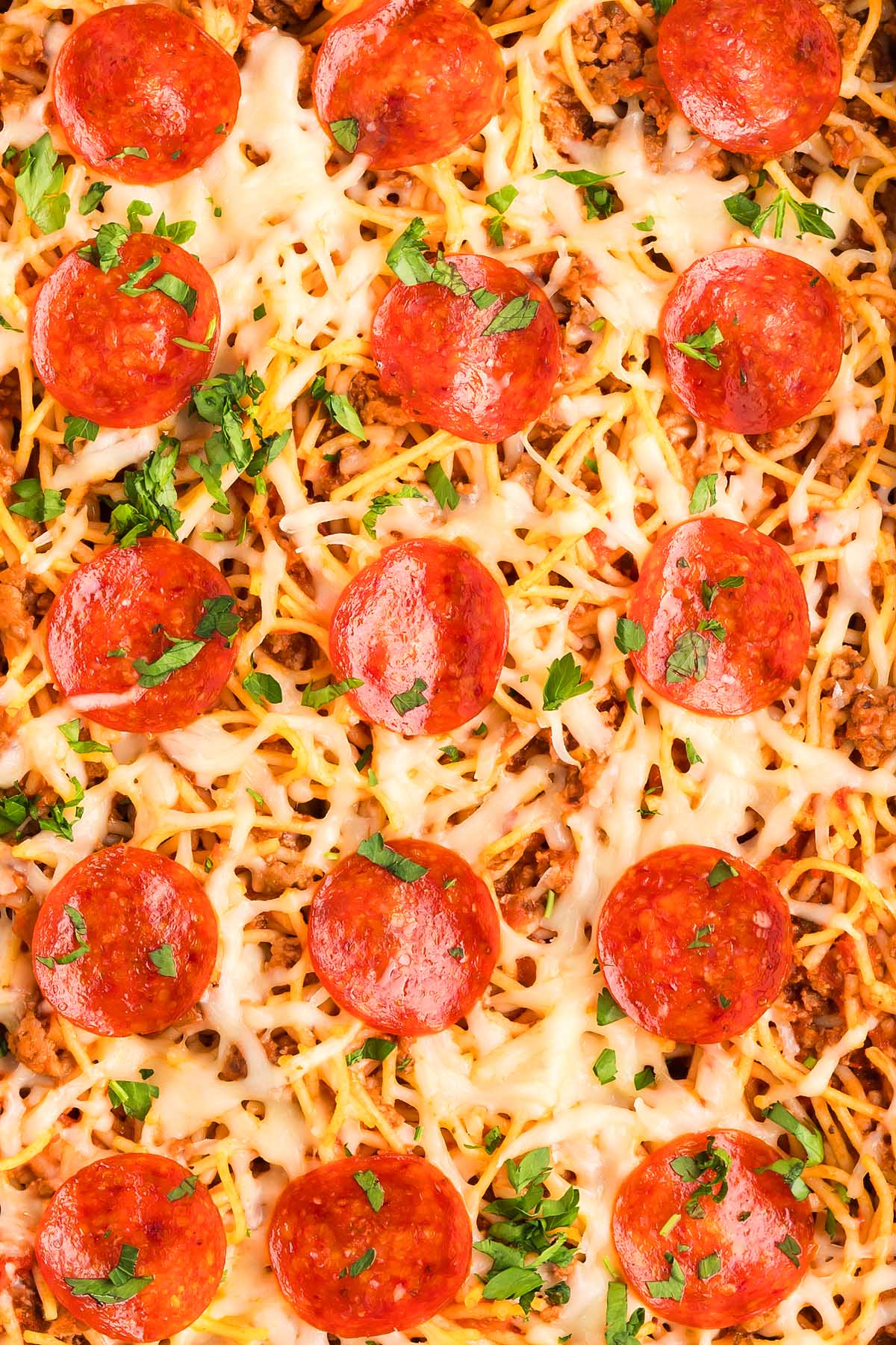 A close-up of baked spaghetti pizza, topped with melted cheese, pepperoni slices, and chopped parsley.