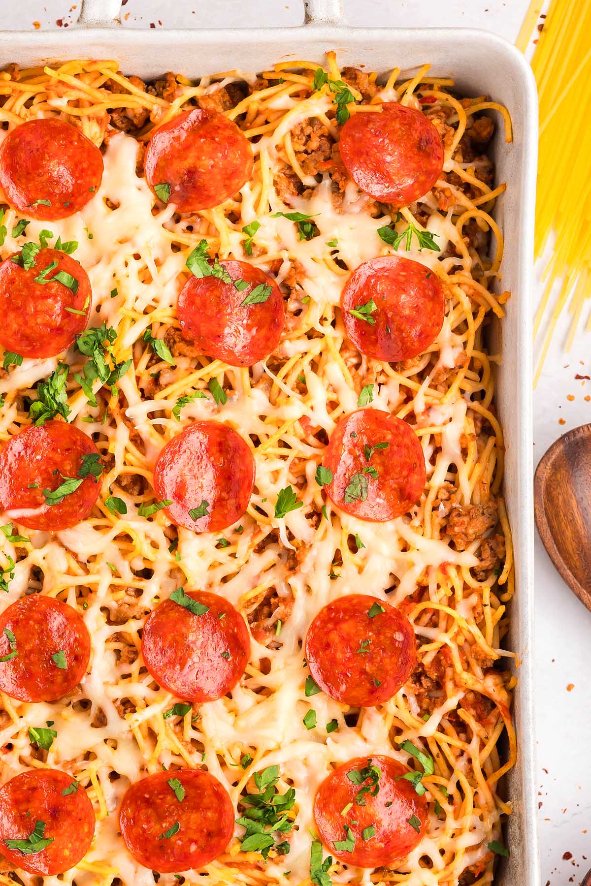 Baked spaghetti pizza topped with melted cheese, pepperoni slices, and chopped parsley in a rectangular dish.