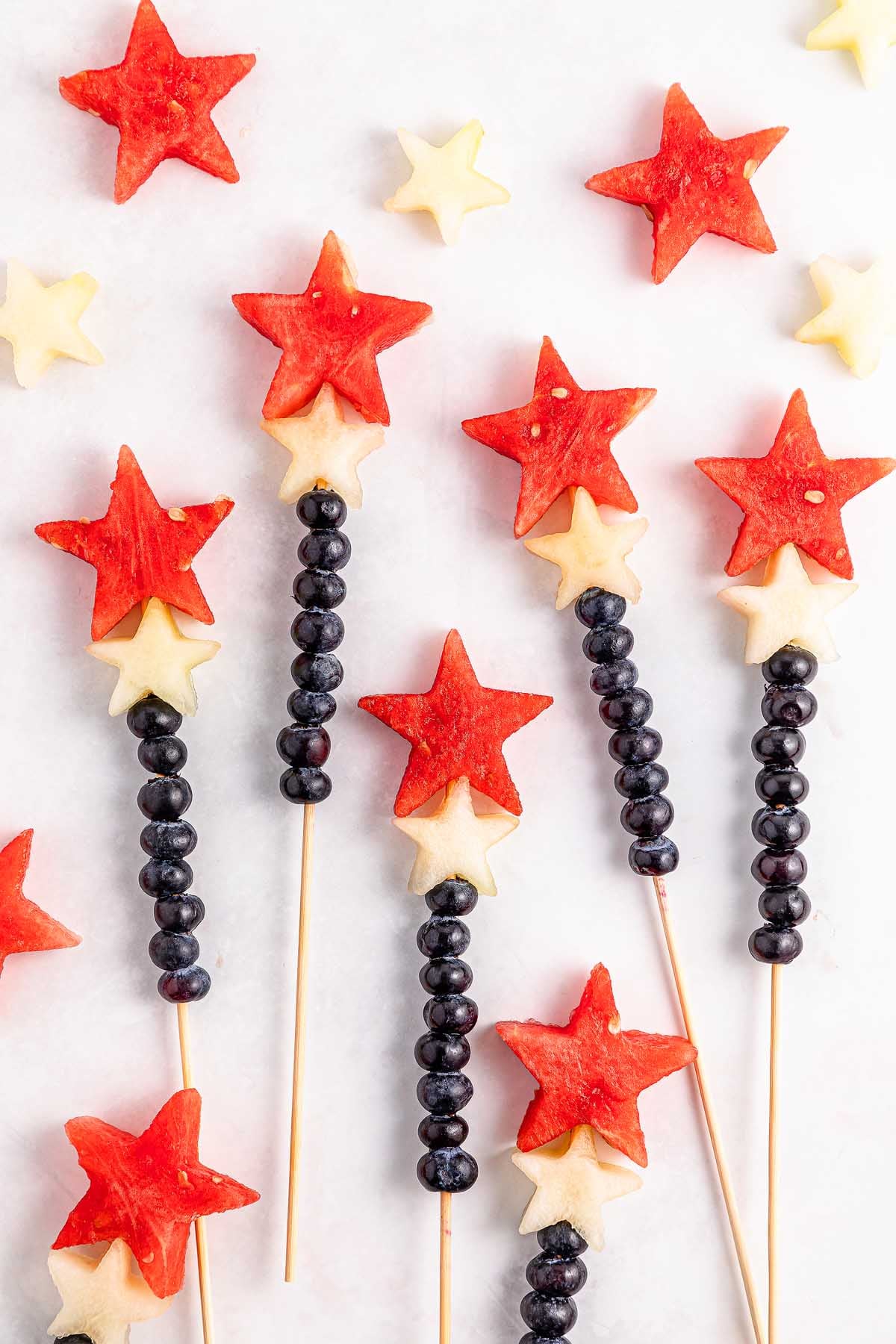Watermelon Blueberry Sparklers: Fruit kabobs featuring watermelon and melon stars with blueberries on skewers, arranged on a white background.