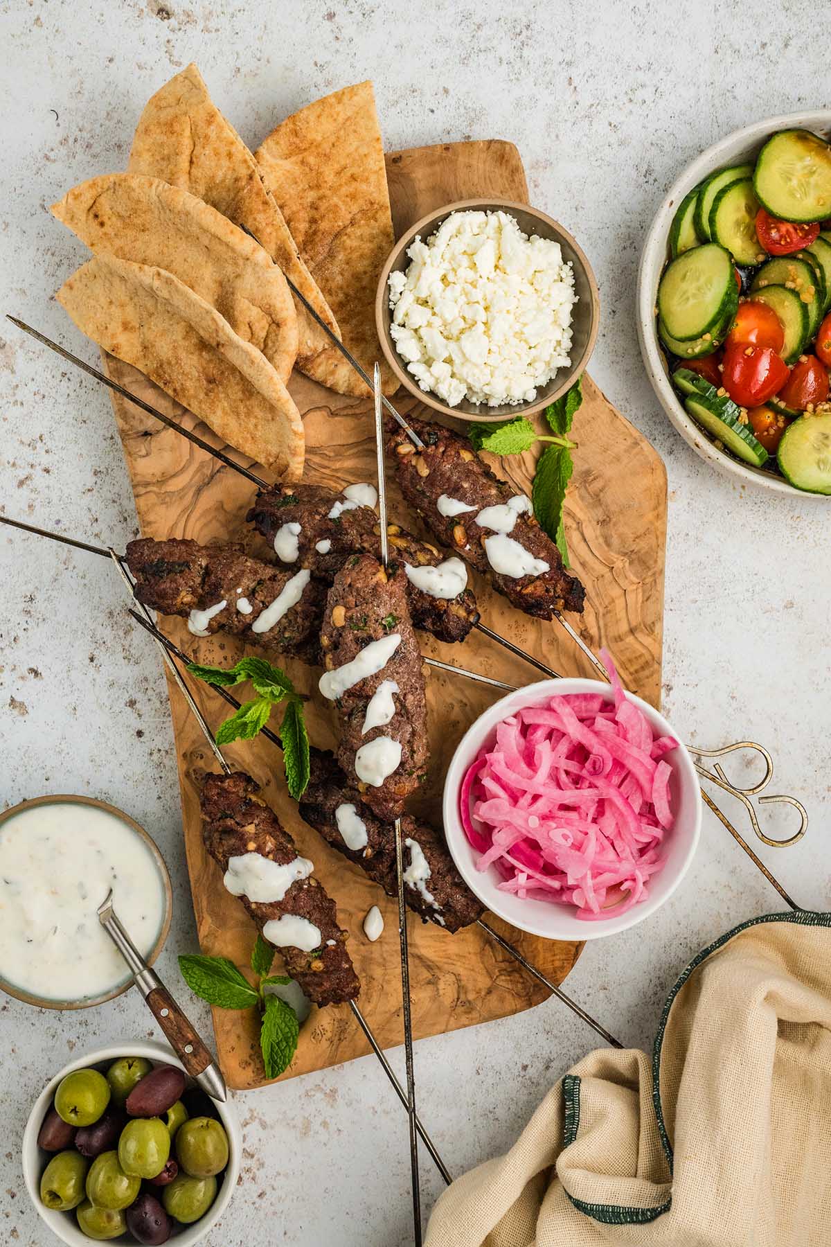 Grilled beef kofta skewers with sauce, pita bread, feta, pickled onions, olives, and salad on a wooden board.