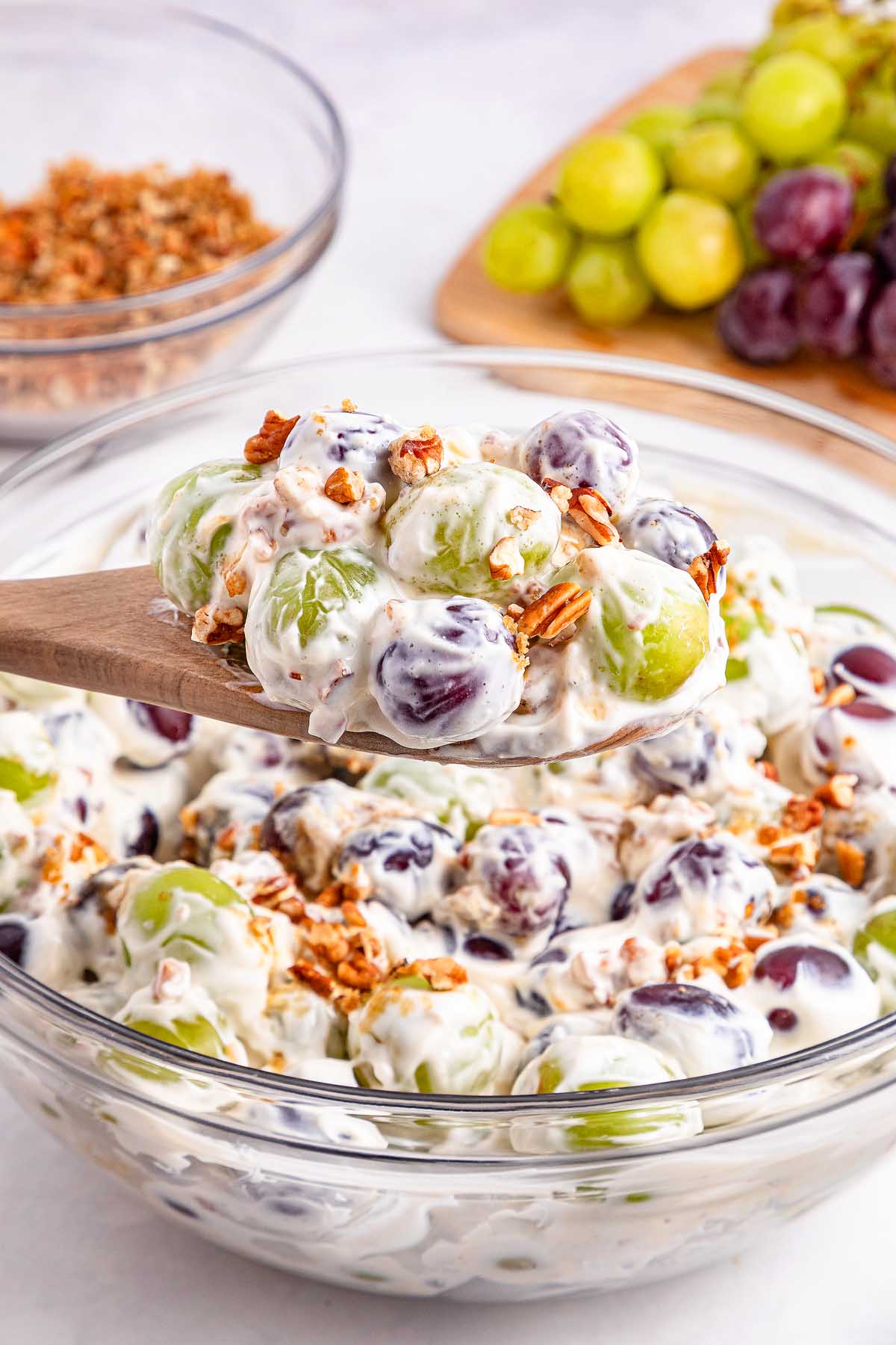 A wooden spoon holds creamy grape salad with granola above a glass bowl; fresh grapes and crunchy granola sit nearby.