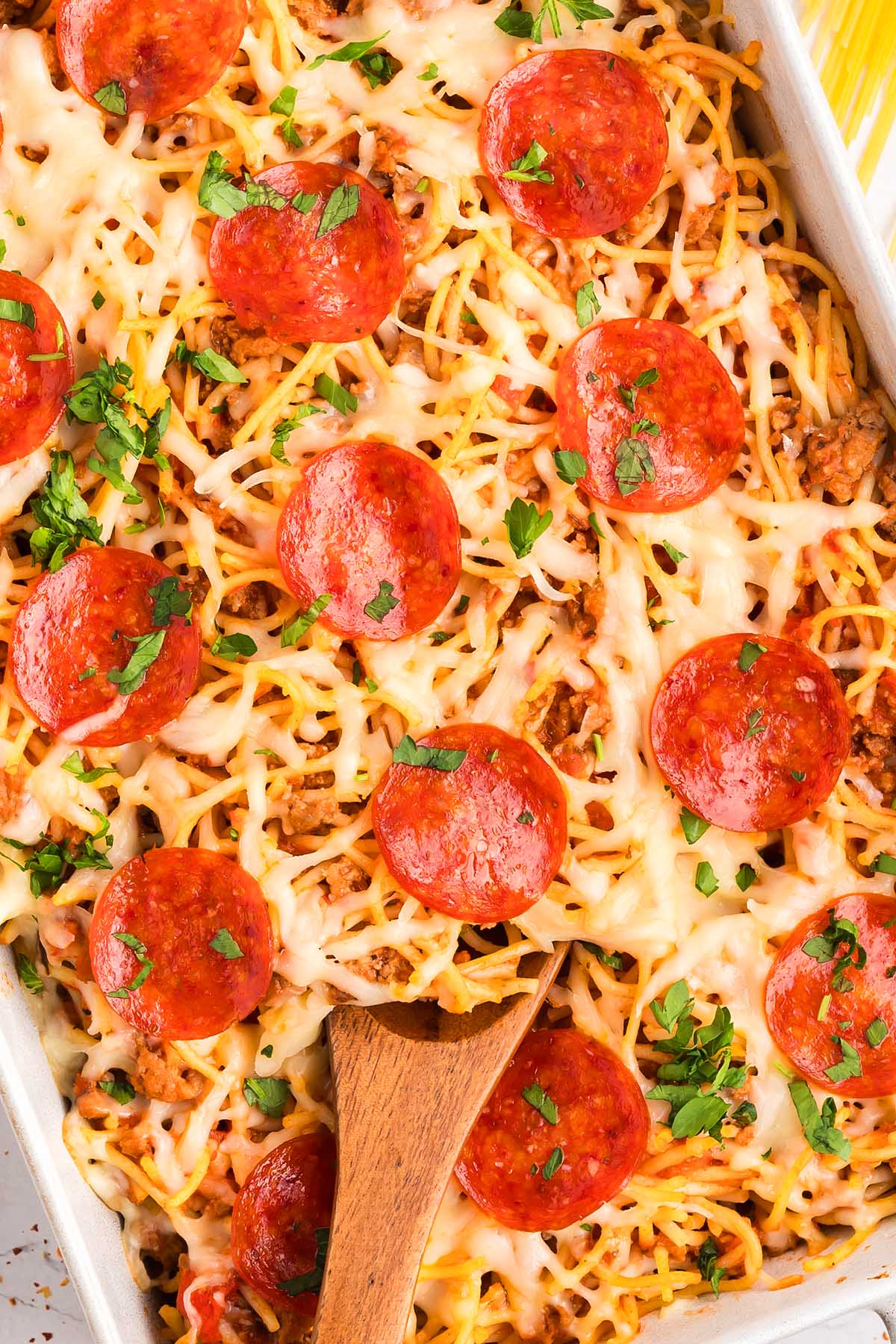 Baked spaghetti pizza casserole topped with melted cheese, pepperoni slices, and chopped herbs, with a wooden spoon in it.