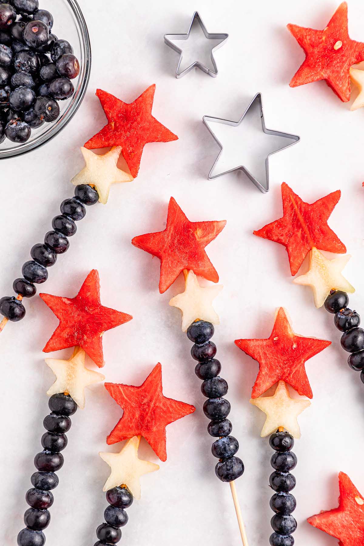 Watermelon Blueberry Sparklers: Fruit skewers with star-shaped watermelon and melon pieces, plus blueberries, arranged on a white surface.