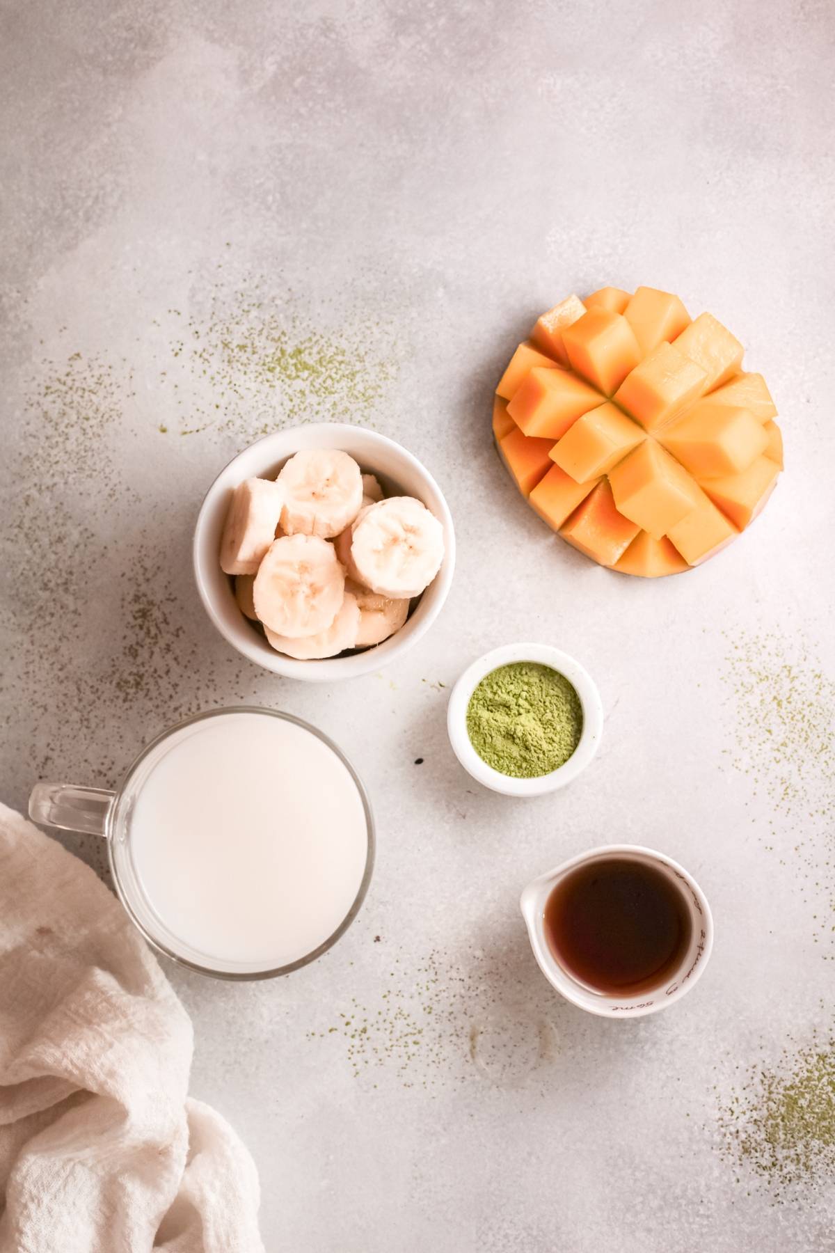 Sliced bananas, cubed mango, milk, green powder, and dark liquid in cups on a light surface—perfect ingredients for a refreshing Mango Matcha Smoothie or your favorite vegan smoothie recipe.