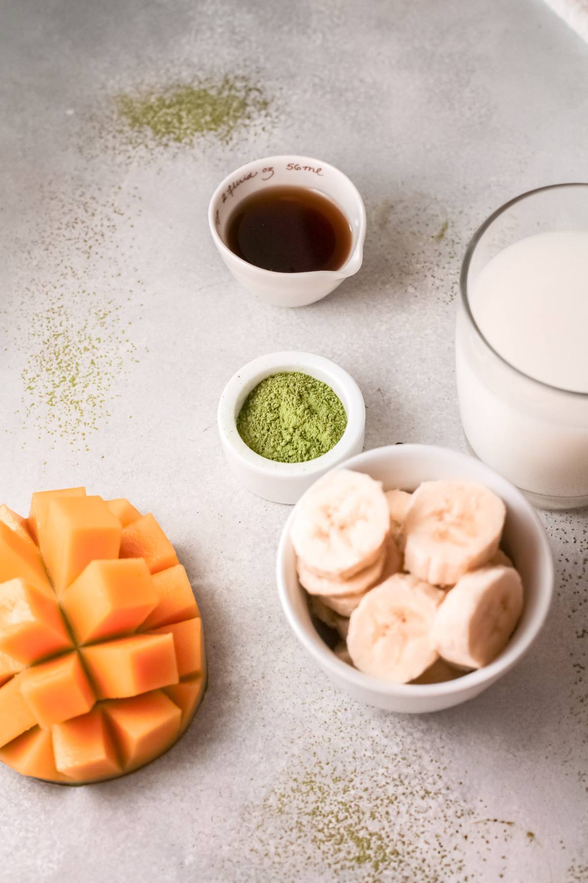 Sliced mango, banana, matcha powder, a cup of milk, and a small cup of liquid on a light surface—perfect ingredients for a refreshing green smoothie with almond milk.