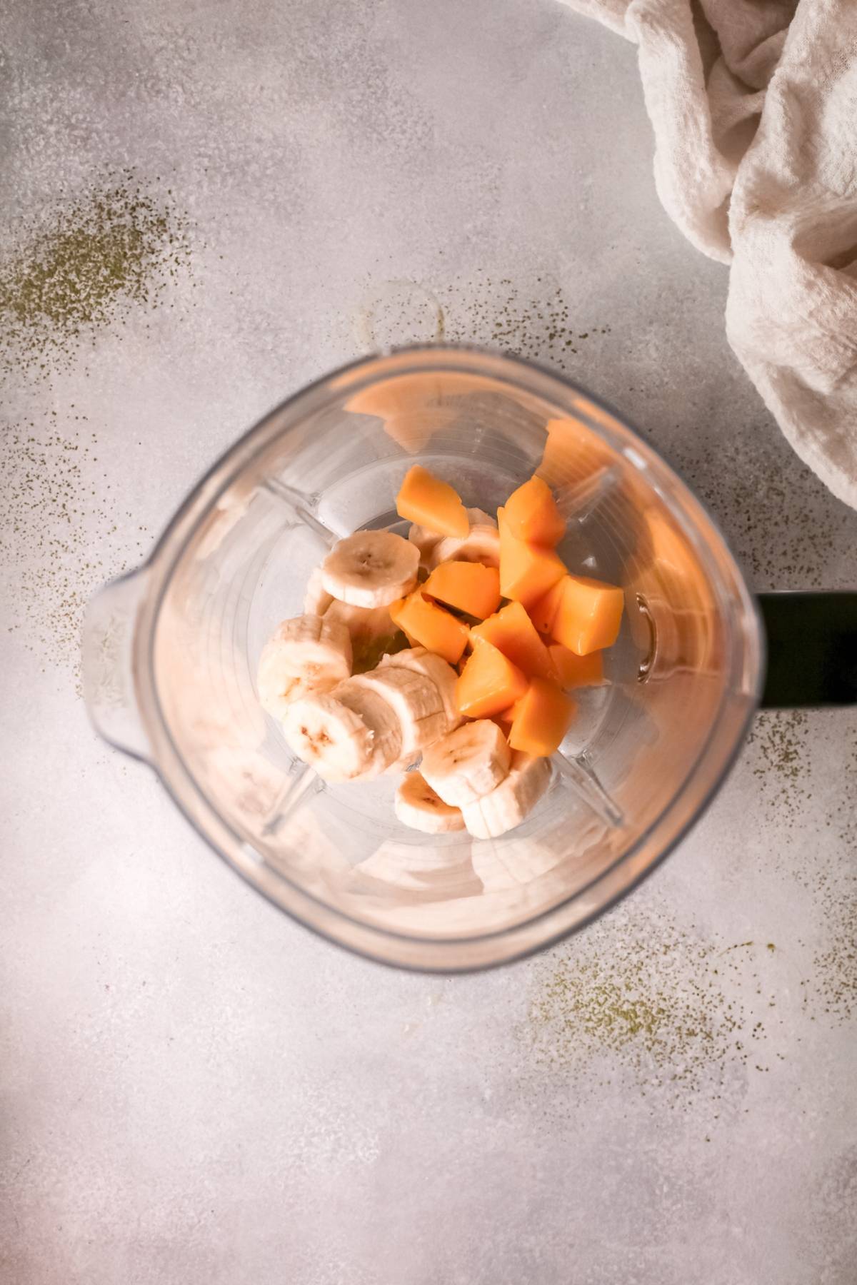 Top view of a blender with sliced banana and cubed papaya inside, ready to be blended into a refreshing green mango smoothie, on a light countertop.