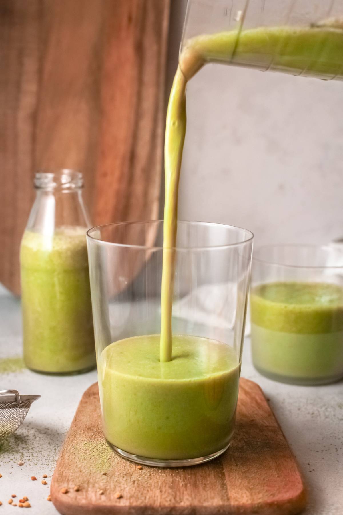 A green smoothie with almond milk is being poured into a glass, with bottles and glasses filled with the vibrant green smoothie visible in the background.