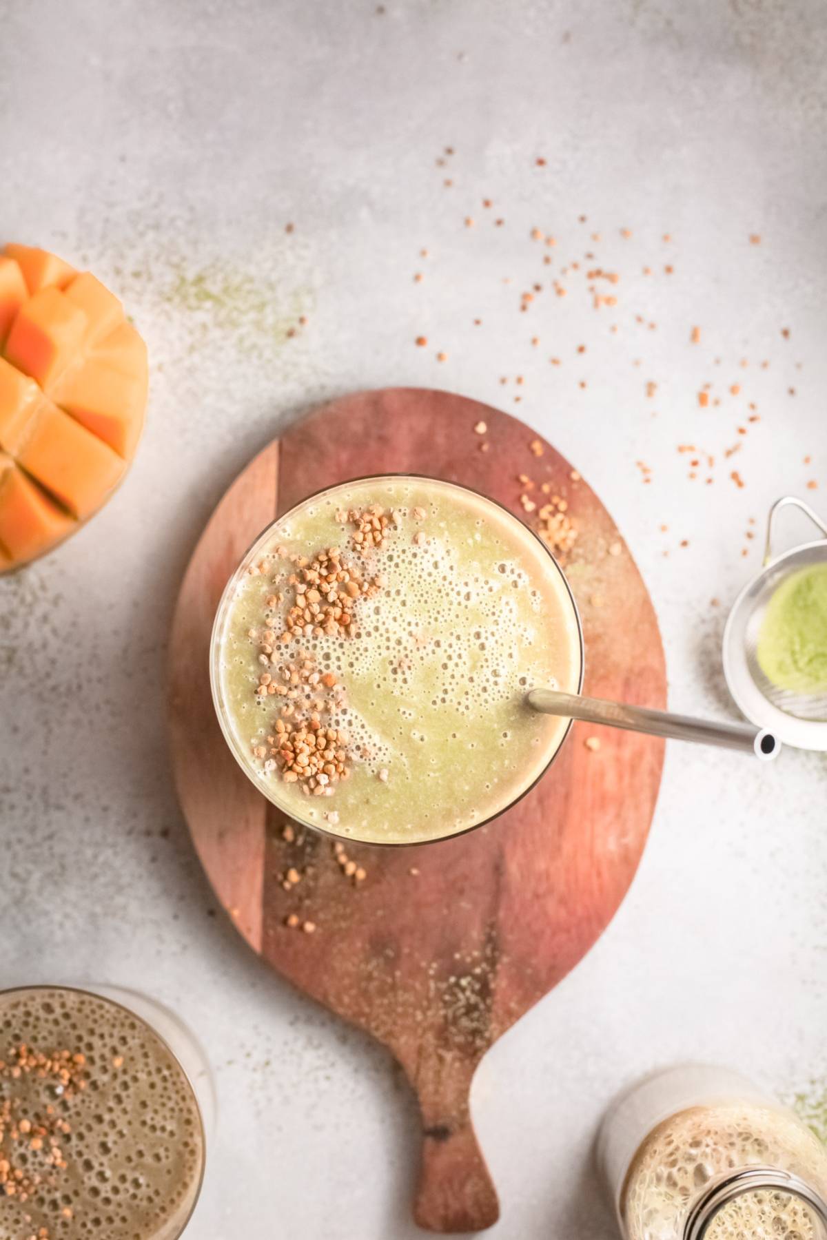 A glass of green mango smoothie topped with seeds sits on a wooden board, next to sliced mango and small bowls—an inviting vegan smoothie recipe.